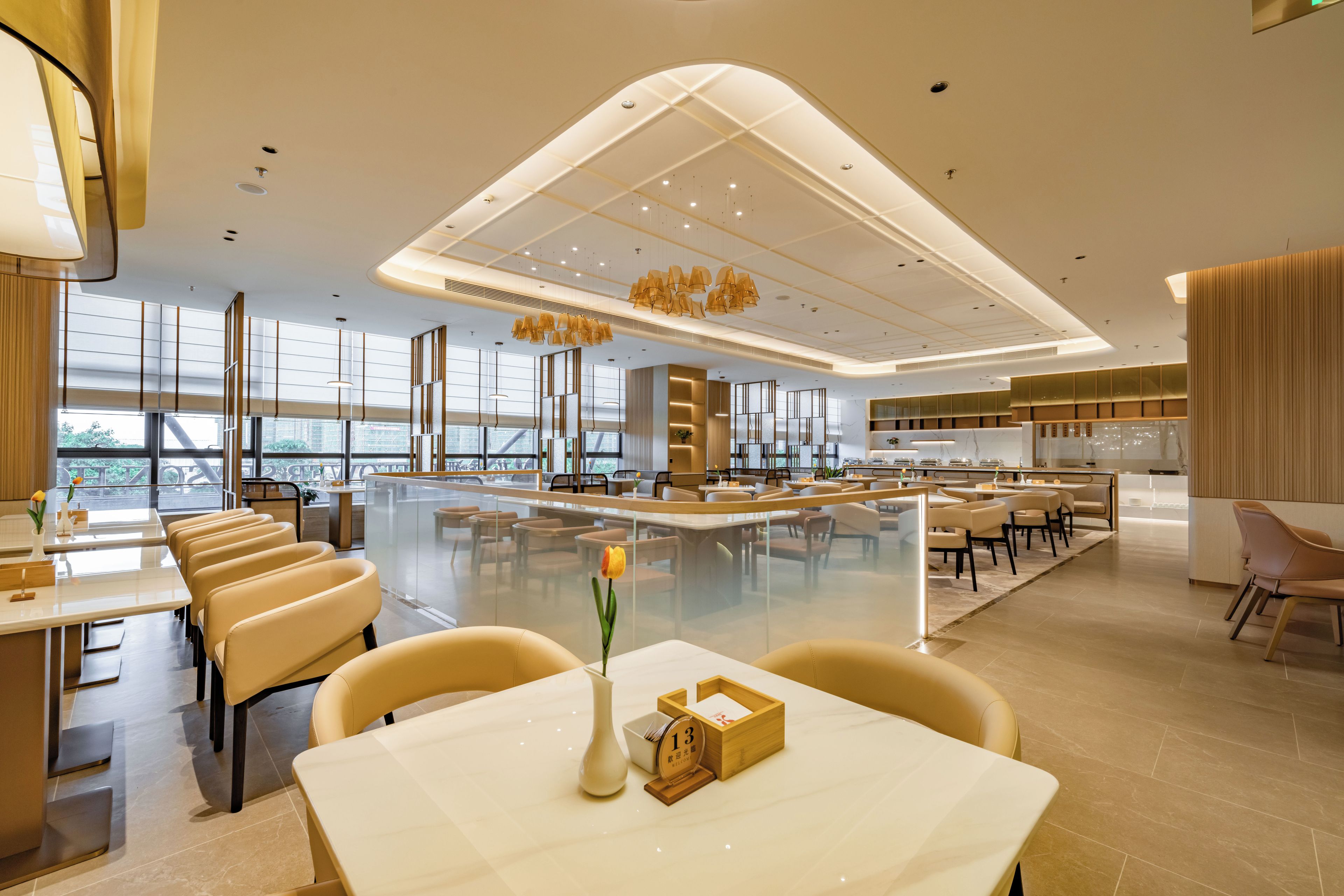 Un amplio comedor de hotel con varias mesas y sillas, ventanas grandes y luminarias modernas.