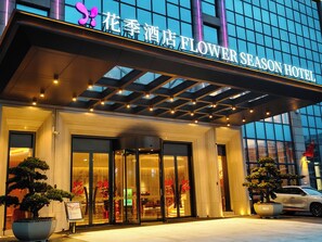 Entrada de hotel com fachada de vidro, placa com a inscrição 'Flower Season Hotel' e um carro estacionado em frente.
