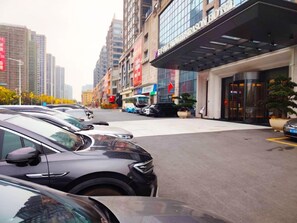 A hotel entrance with a modern design, a parking lot with several cars, and high-rise buildings in the background.