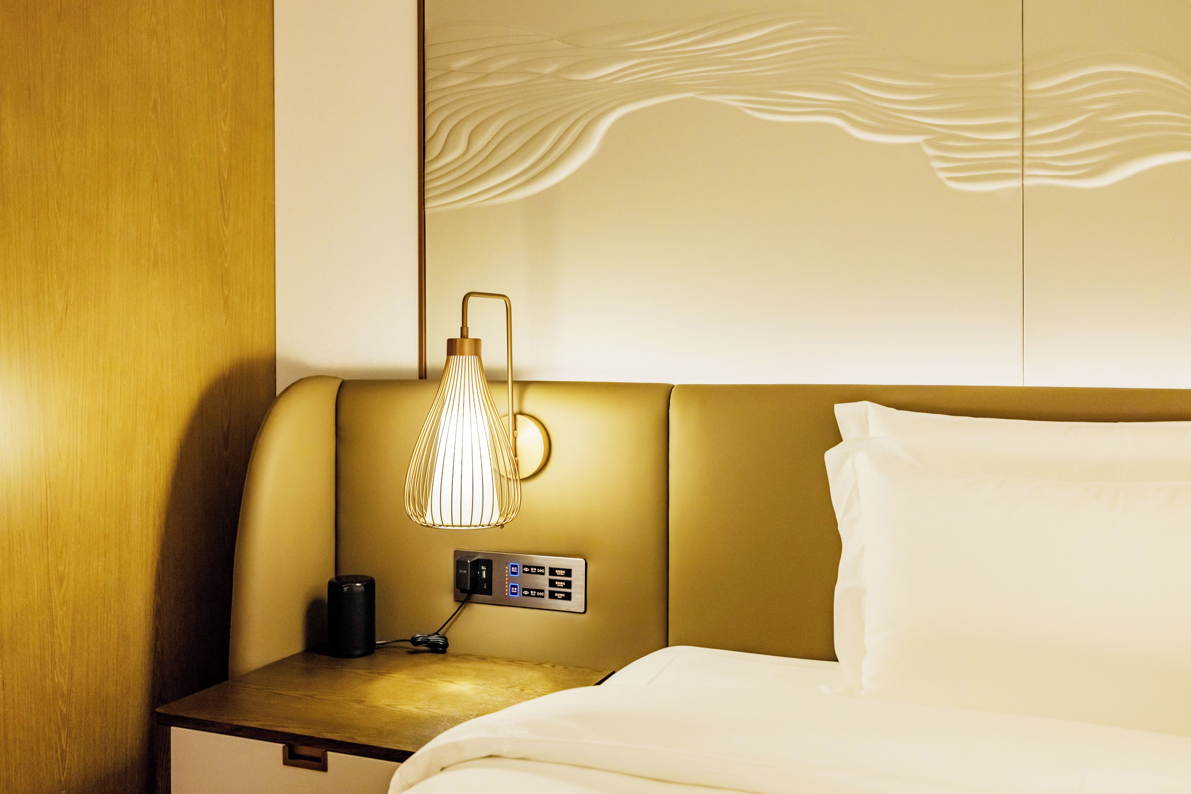A hotel room with a bed, a bedside table with a lamp, and a wall panel.