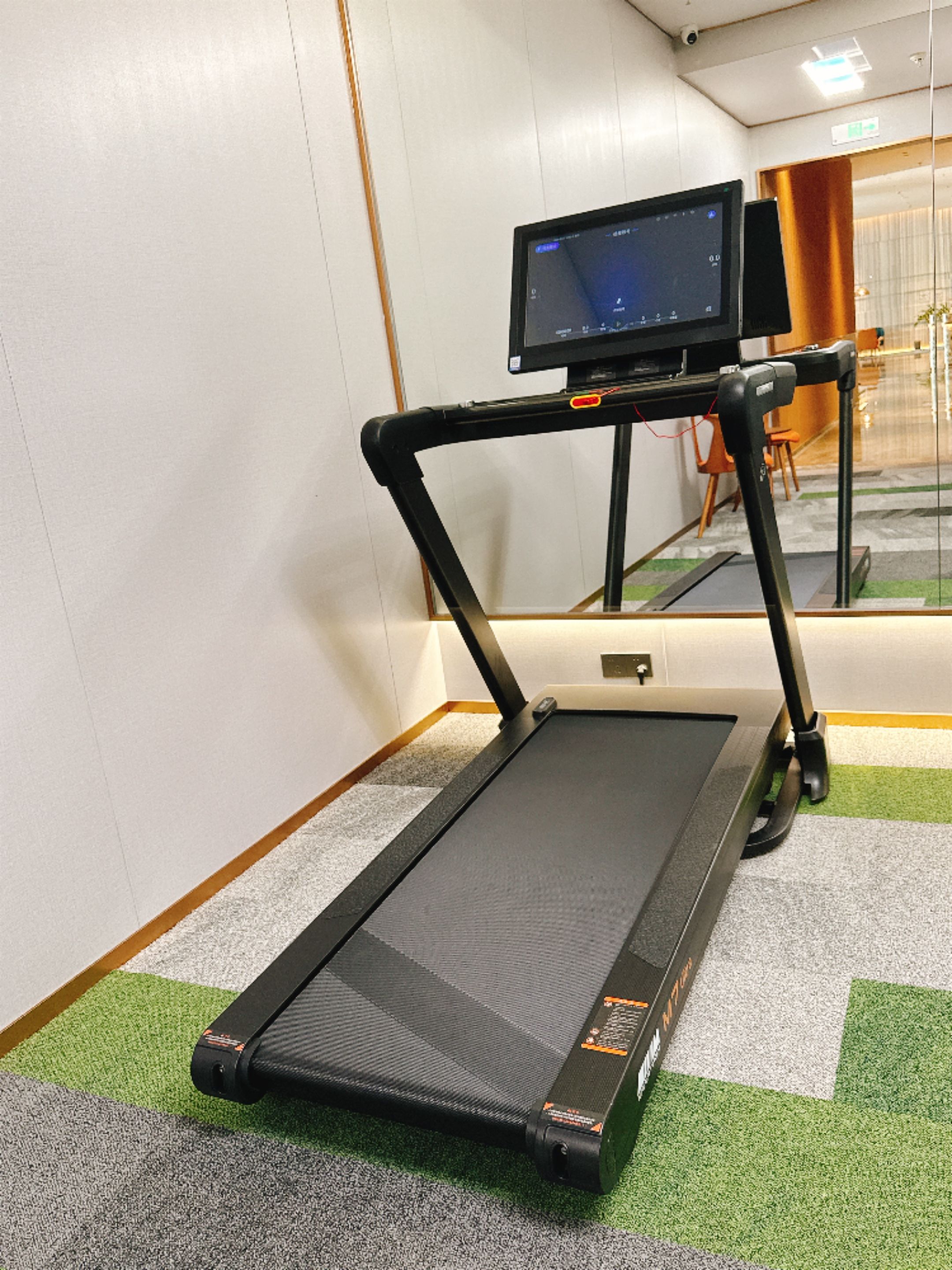Una cinta de correr en una habitación con piso alfombrado, un monitor sobre un pedestal, y el reflejo de una silla en el espejo.