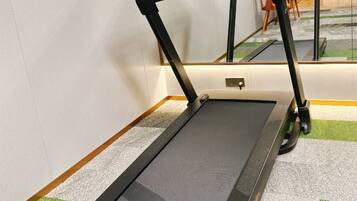Una cinta de correr en una habitación con piso alfombrado, un monitor sobre un pedestal, y el reflejo de una silla en el espejo.