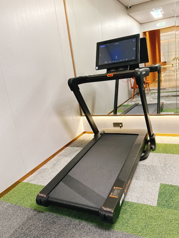 A treadmill in a room with a carpeted floor, a monitor on a stand, and a reflection of a chair in the mirror.