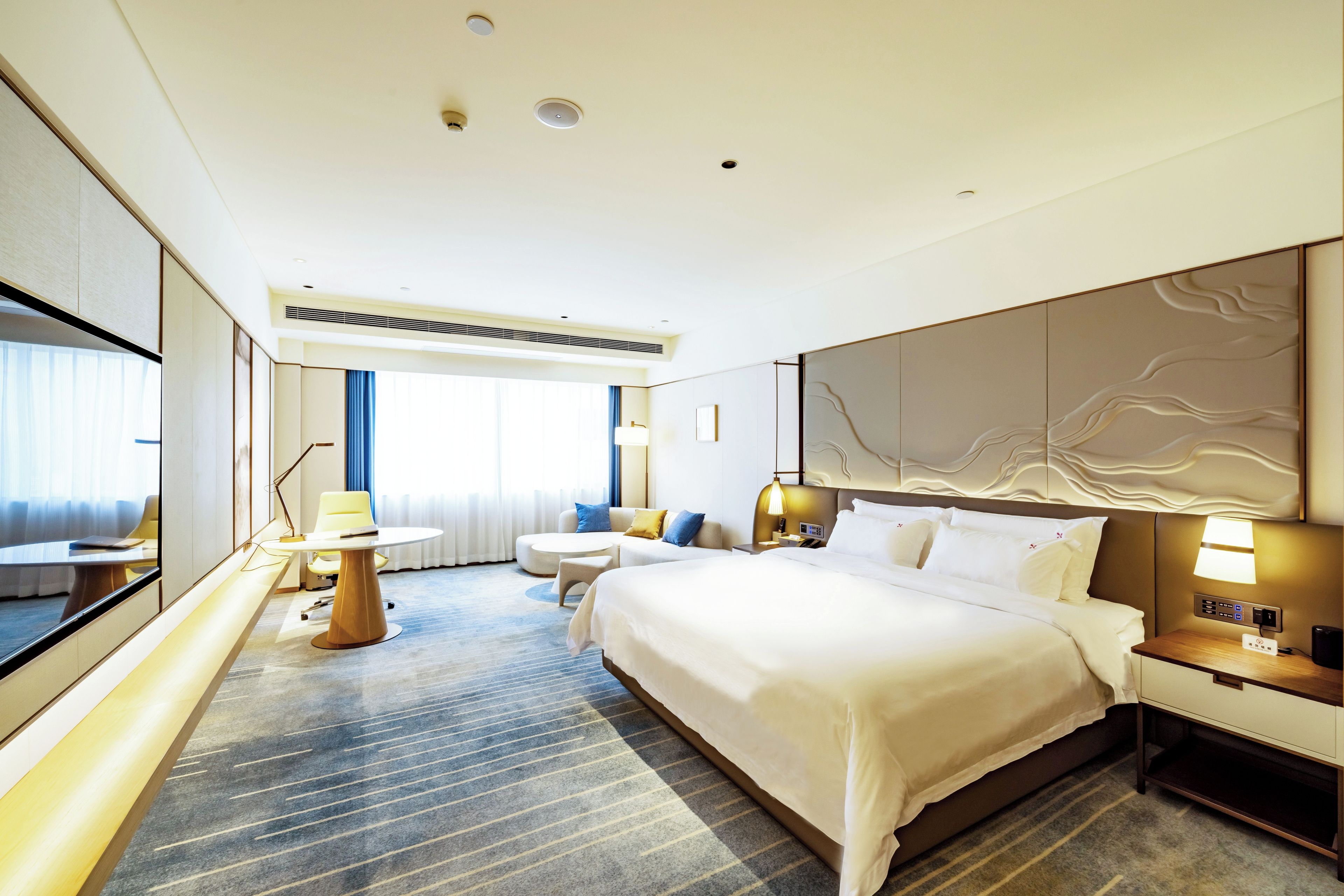 Habitación de hotel con una cama grande, un escritorio, una silla, un sofá, un televisor y un ventanal amplio.