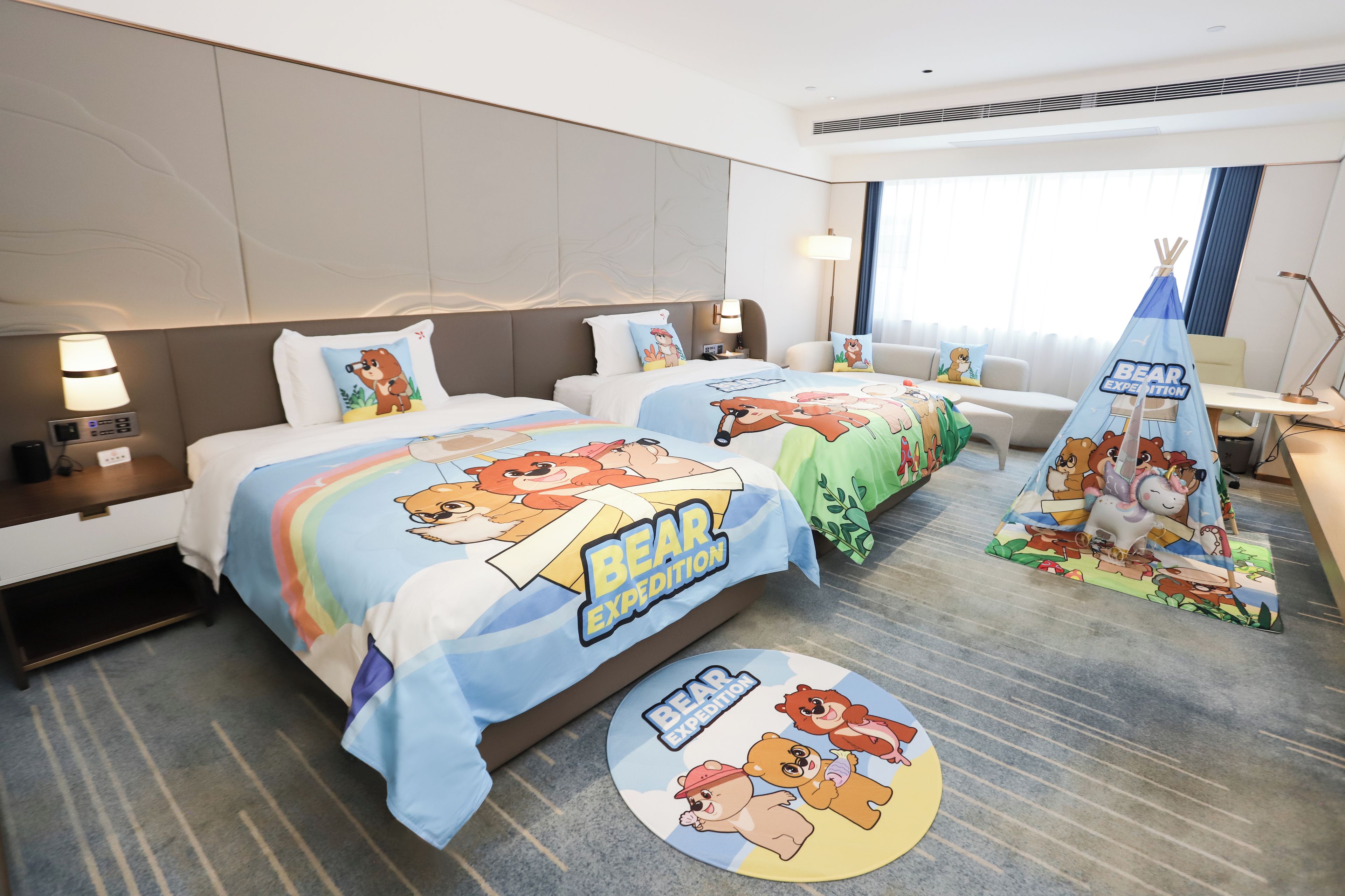 Habitación de hotel con dos camas, cada una con cubrecama con temática de oso, una mesita con lámpara y una decoración de tienda de campaña.