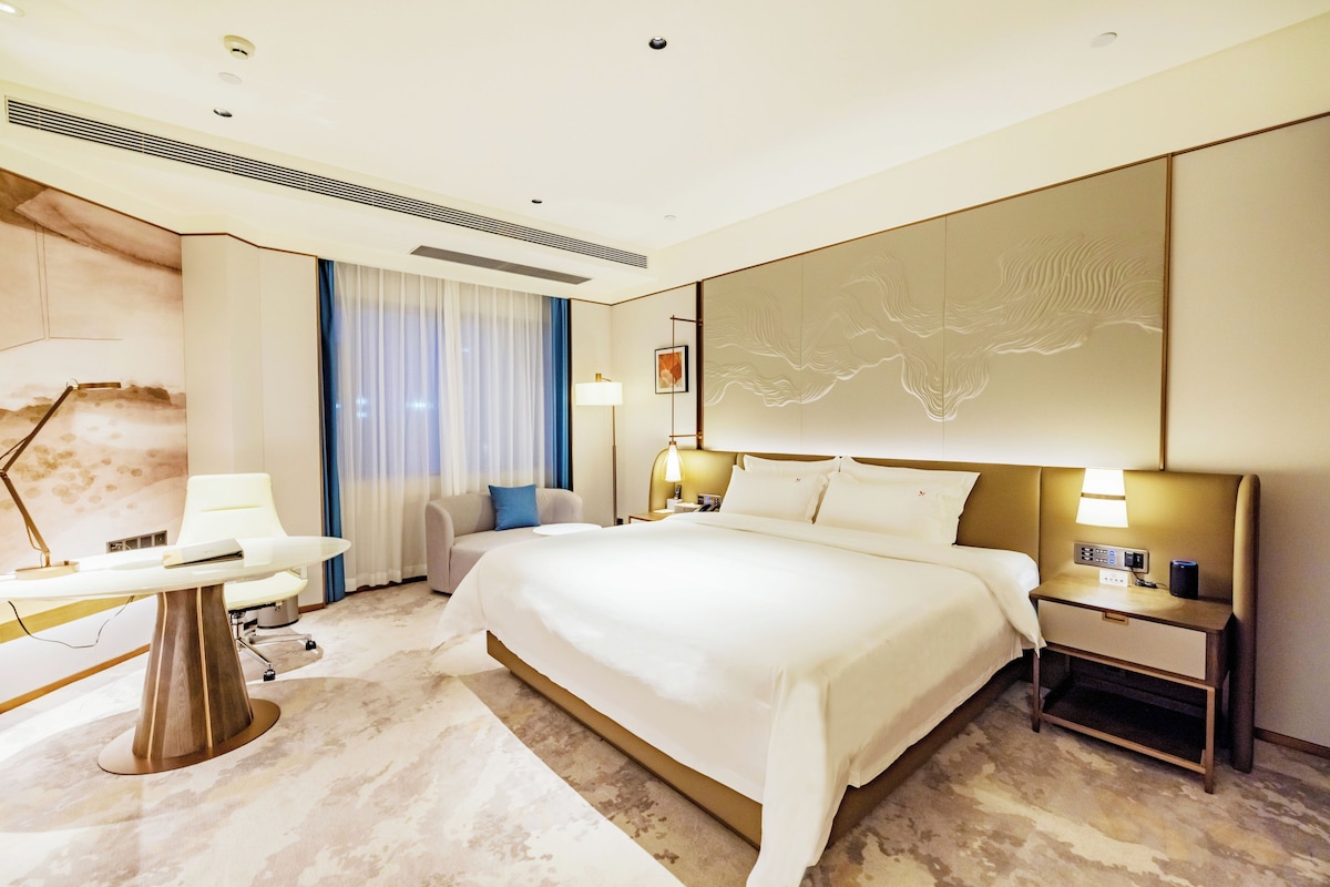Quarto de hotel com uma cama grande, mesas de cabeceira, escrivaninha, uma cadeira, abajur e um painel decorativo na parede.