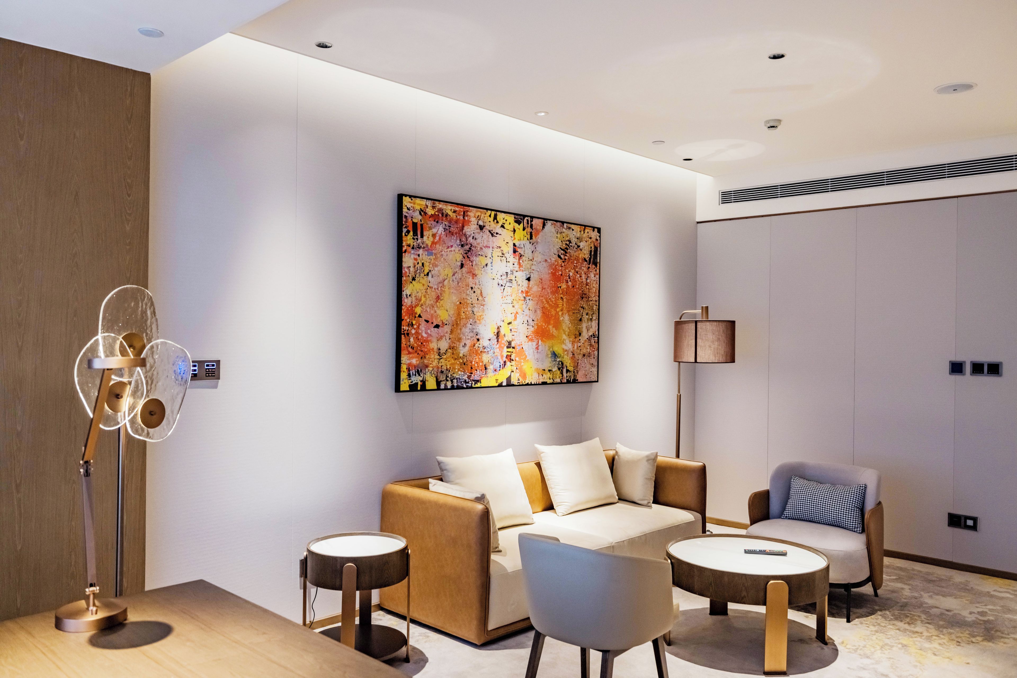 A modern hotel room with a sofa, two armchairs, a coffee table, a floor lamp, and a large abstract painting on the wall.