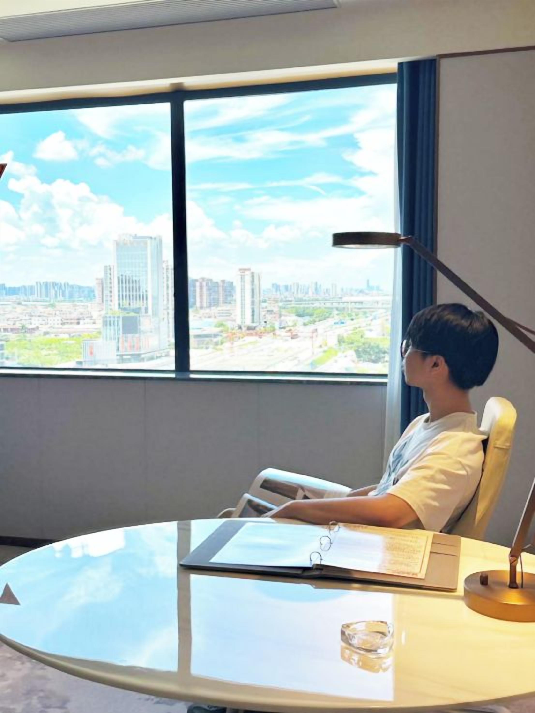 A person seated at a round glass table with a laptop, a notebook, and a pen, in a room with a large window offering a city view.