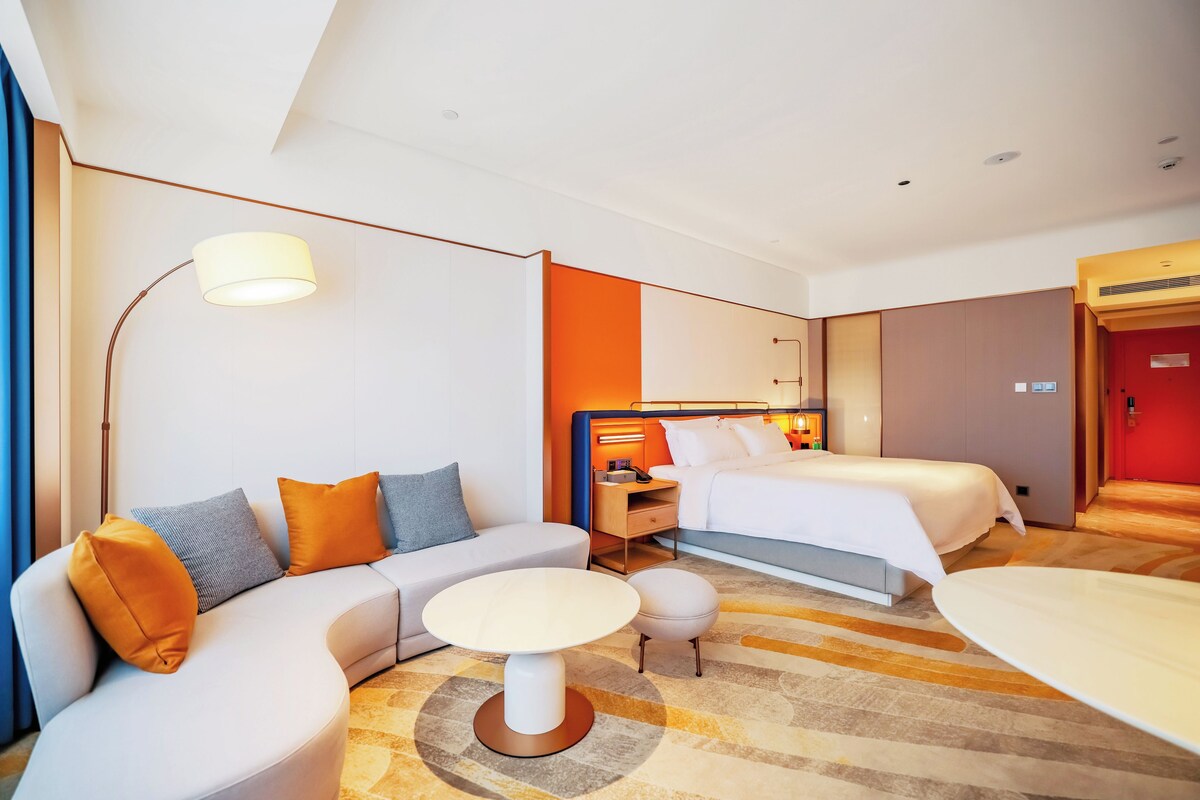 Quarto de hotel com sofá, duas camas, uma mesa pequena e uma luminária.