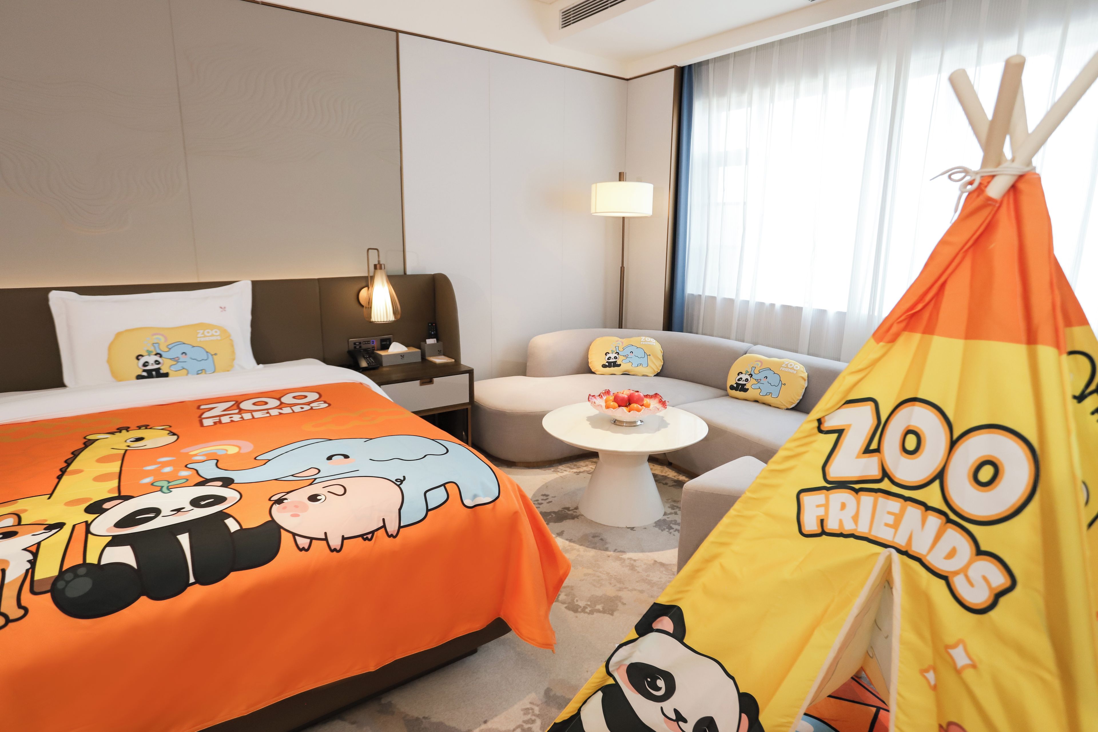 A hotel room with a bed featuring 'Zoo Friends' themed bedding, a small round table with a fruit bowl, a sofa with matching cushions, and a tent with 'Zoo Friends' branding.