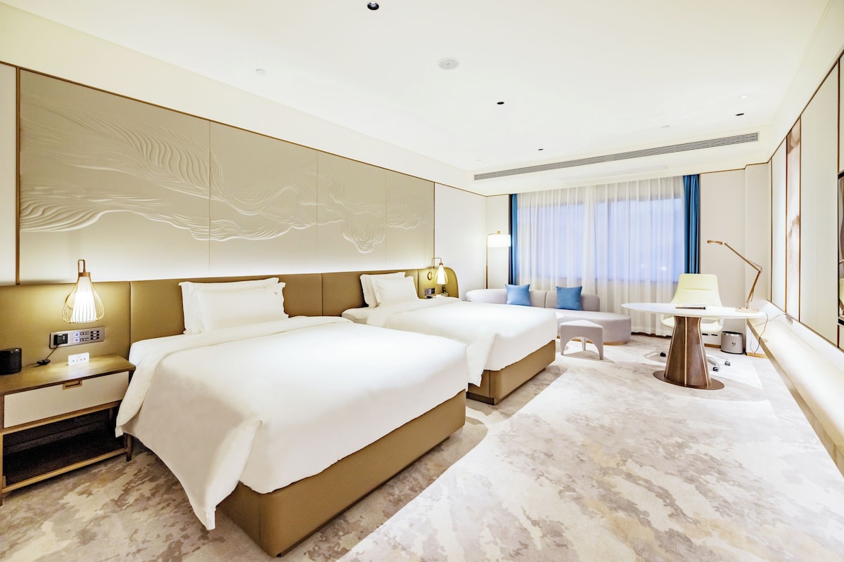 Quarto de hotel com duas camas, uma mesa, uma cadeira, um abajur e uma janela grande com cortinas.