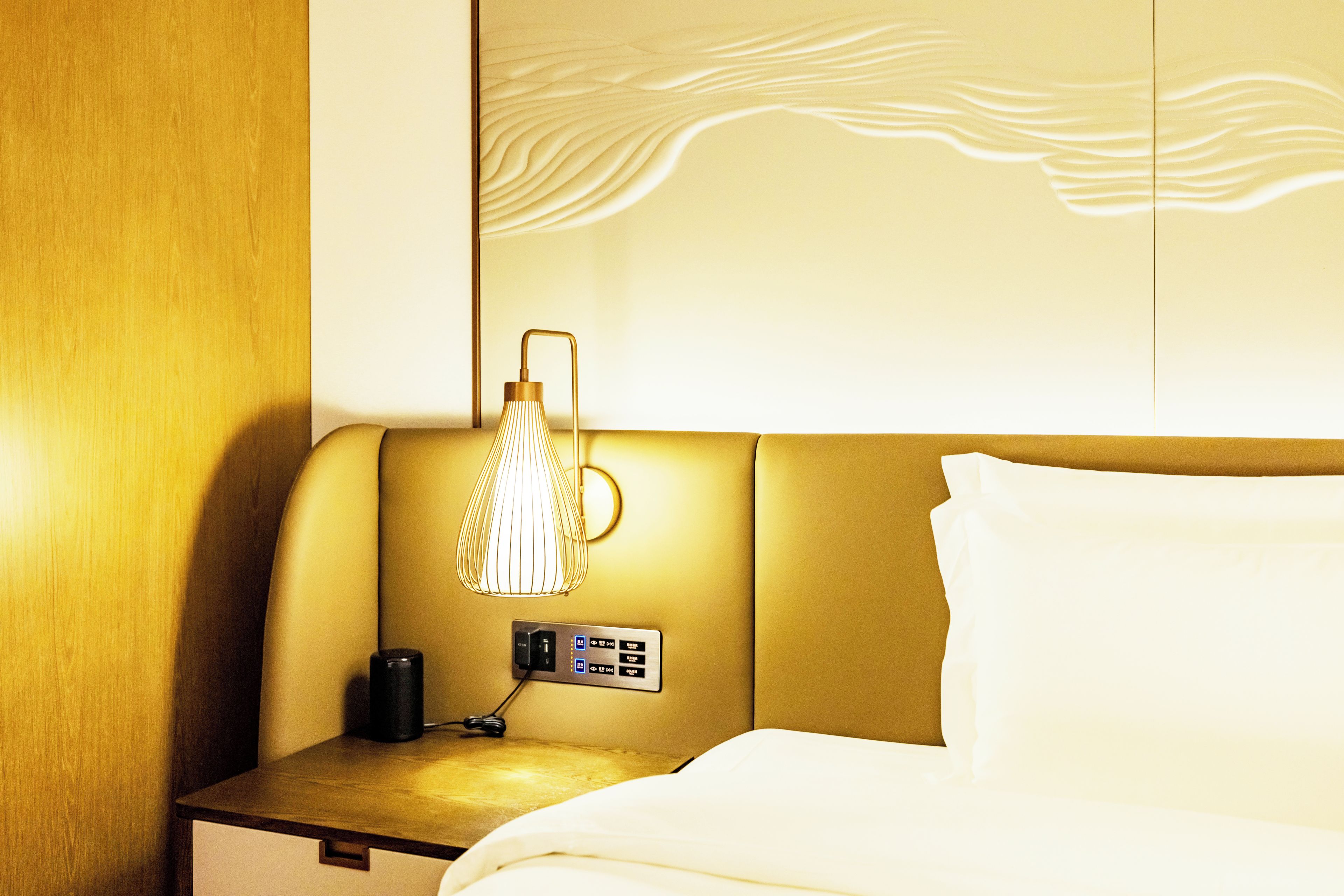 A hotel room with a bed, a nightstand, a lamp, and a wall-mounted control panel.