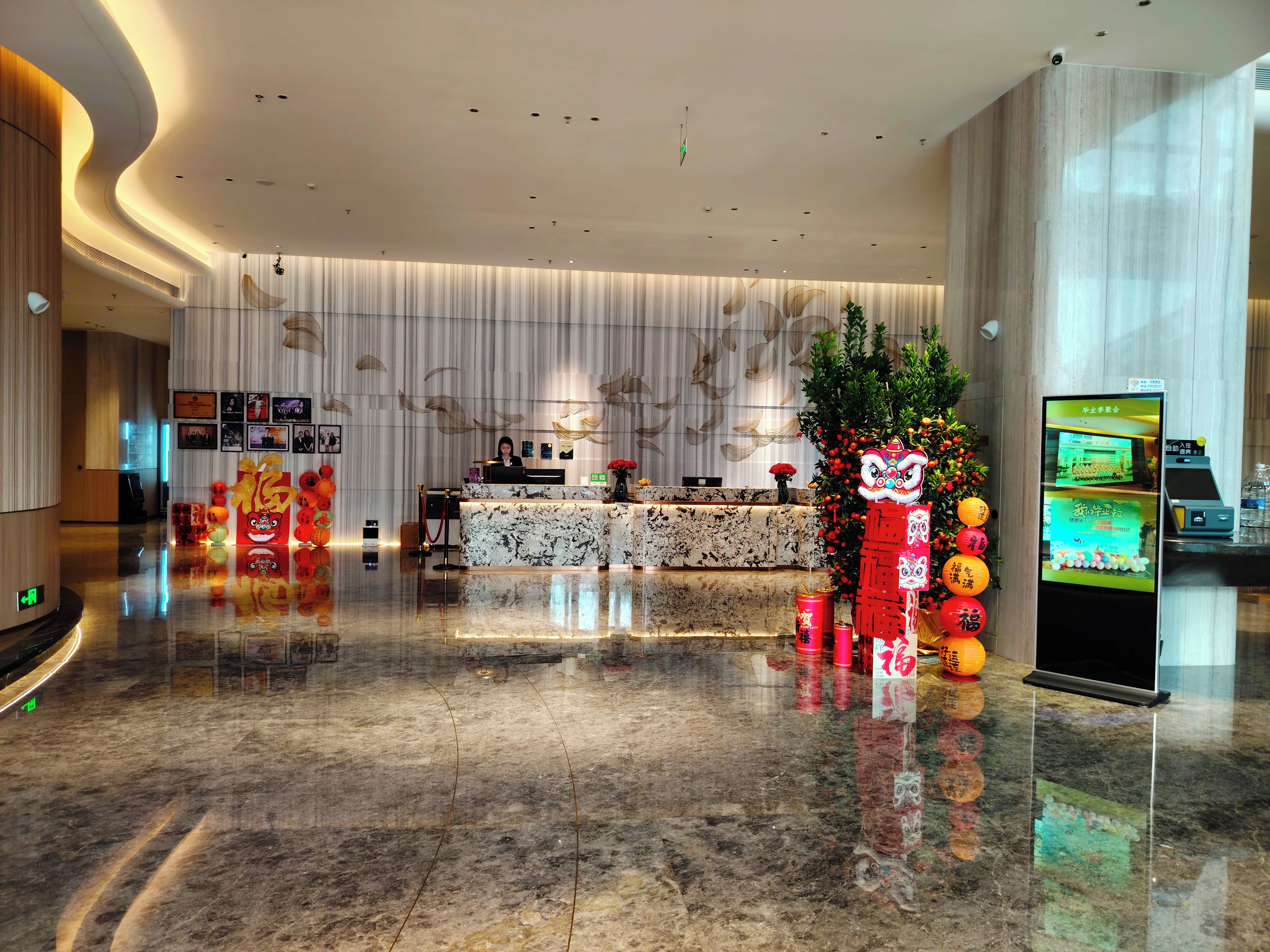A hotel lobby with a reception desk, a digital screen, and a decorative plant.