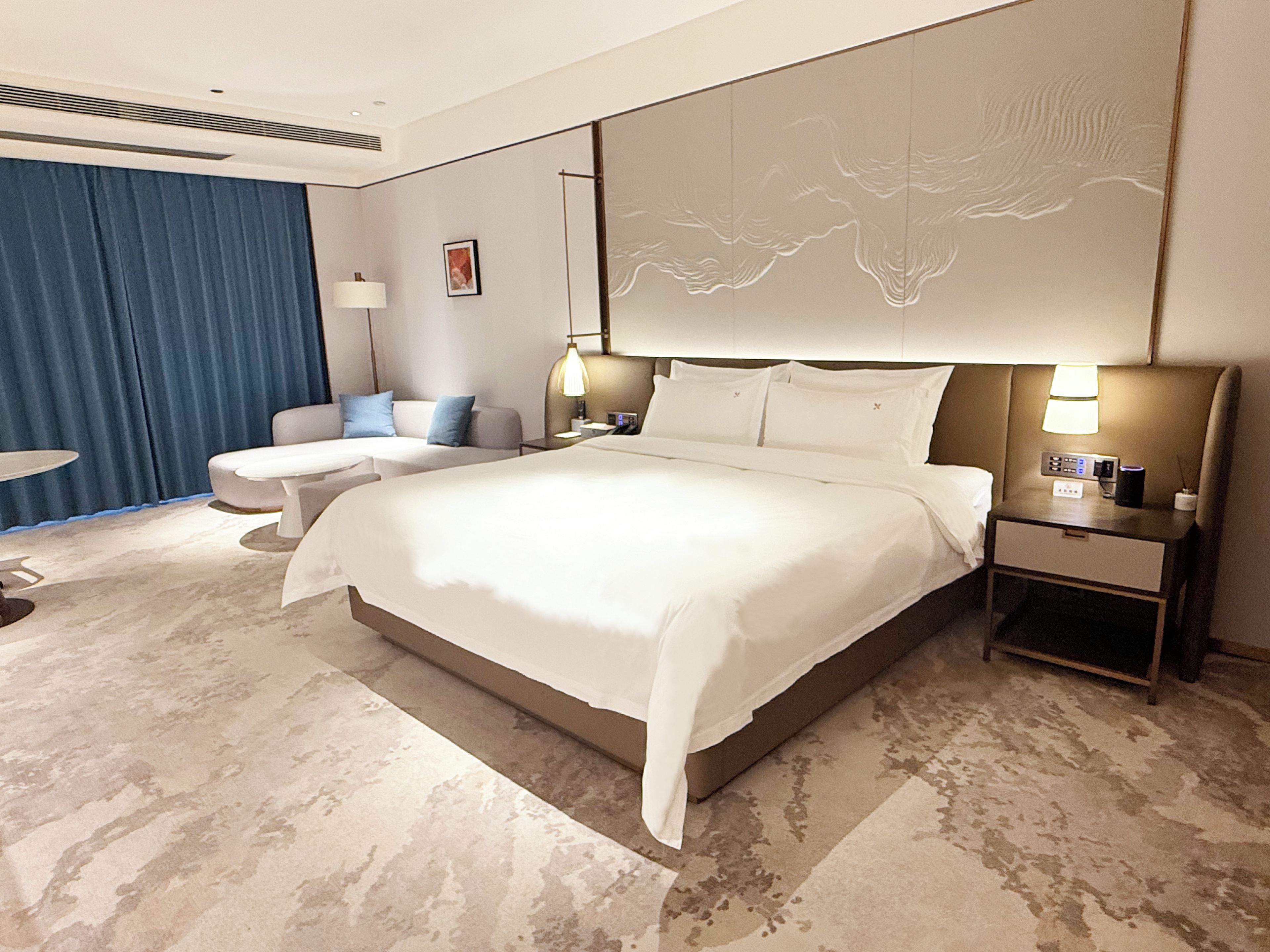 Una habitación de hotel moderna con una cama grande, una zona de estar con un sofá y sillones, una mesita de noche con lámpara y una obra de arte colgada en la pared.