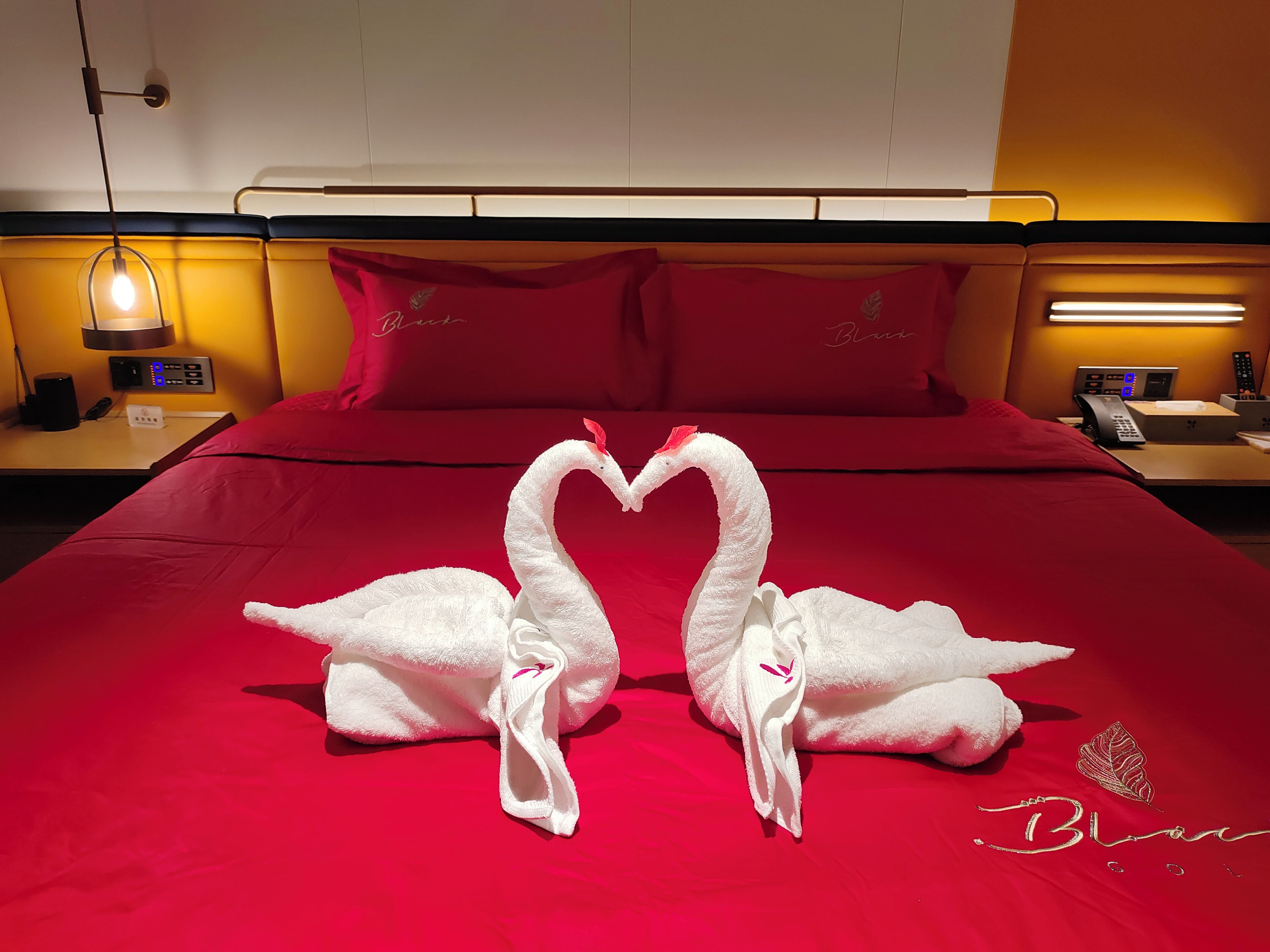 A hotel room with a red bedspread, a white swan towel decoration, a bedside table with a lamp, and a control panel.