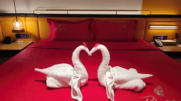 A hotel room with a red bedspread, a white swan towel decoration, a bedside table with a lamp, and a control panel.
