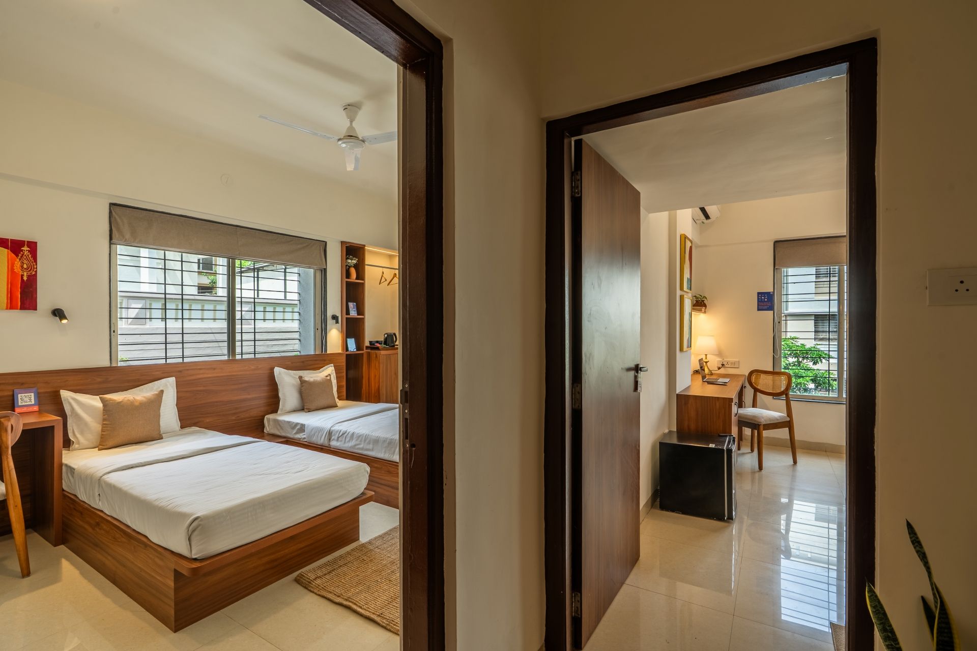 A hotel room with two beds, a desk, a chair, and a small table. There is a window with blinds and a ceiling fan.