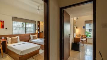 A hotel room with two beds, a desk, a chair, and a small table. There is a window with blinds and a ceiling fan.