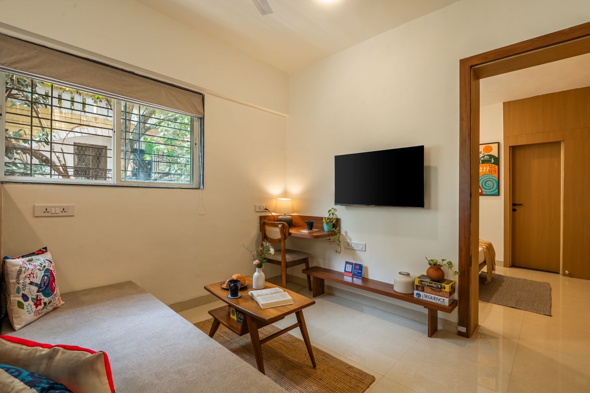 A living room with a sofa, a coffee table, a TV mounted on the wall, and a window with a view of greenery.