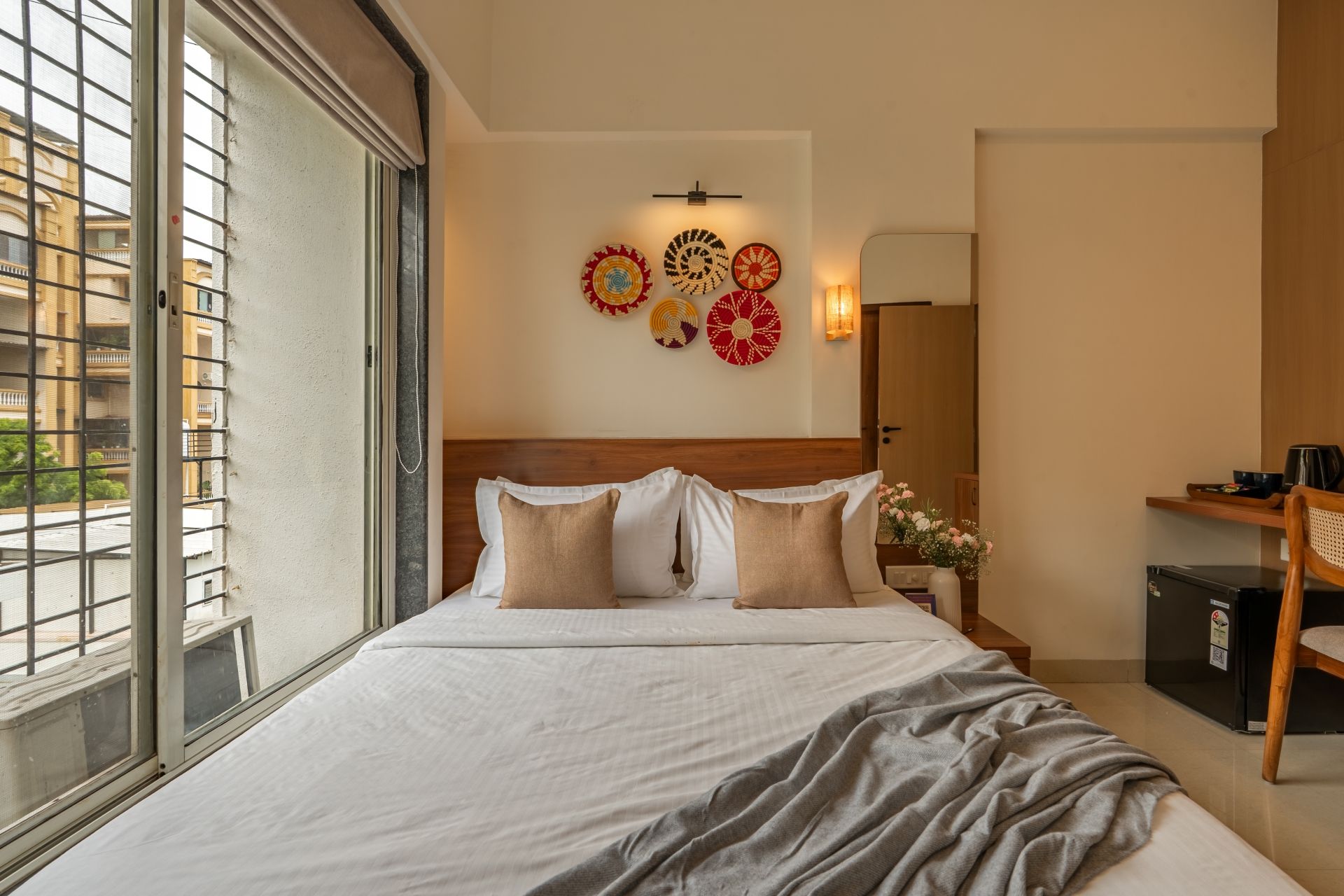 A hotel room with a large bed, a bedside table, a nightstand, a mirror, and a window with a view of buildings.