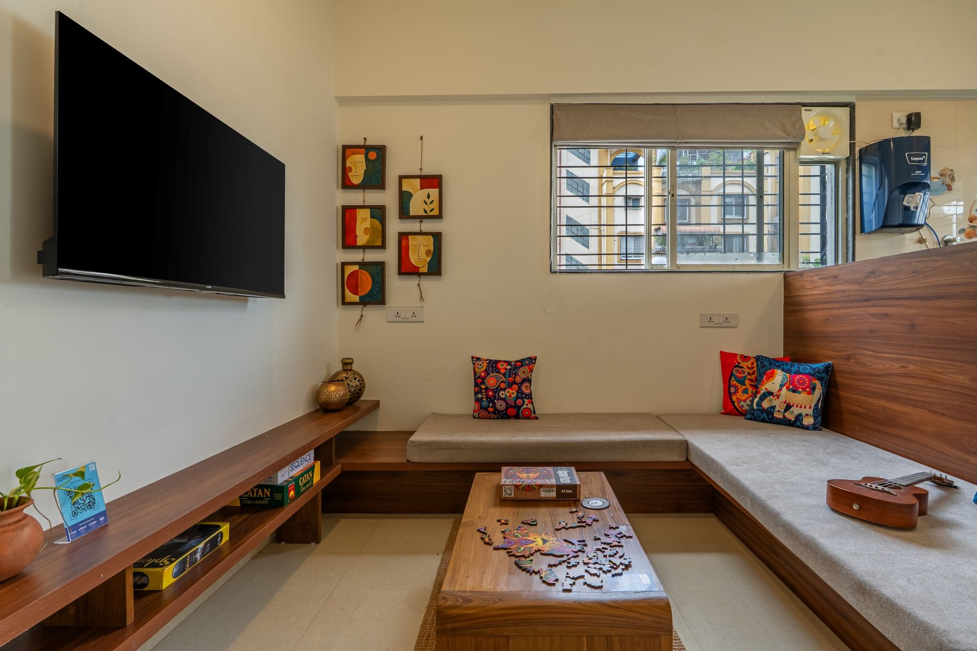 A living room with a flat-screen TV, a wooden coffee table with a puzzle, a sofa with colorful cushions, and a window with a view of buildings.