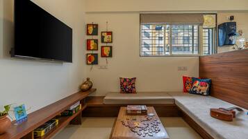 A living room with a flat-screen TV, a wooden coffee table with a puzzle, a sofa with colorful cushions, and a window with a view of buildings.