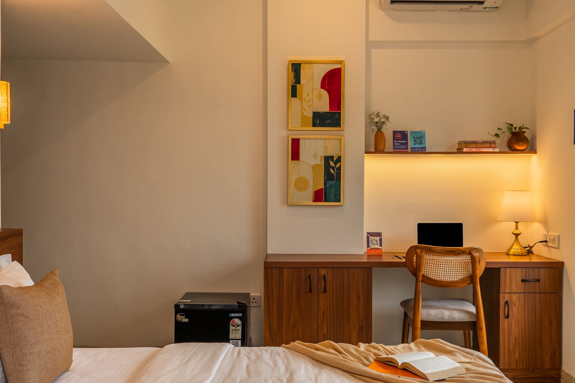 Ett hotellrum med en säng, ett skrivbord, en stol, ett litet svart kylskåp och konst på väggen.