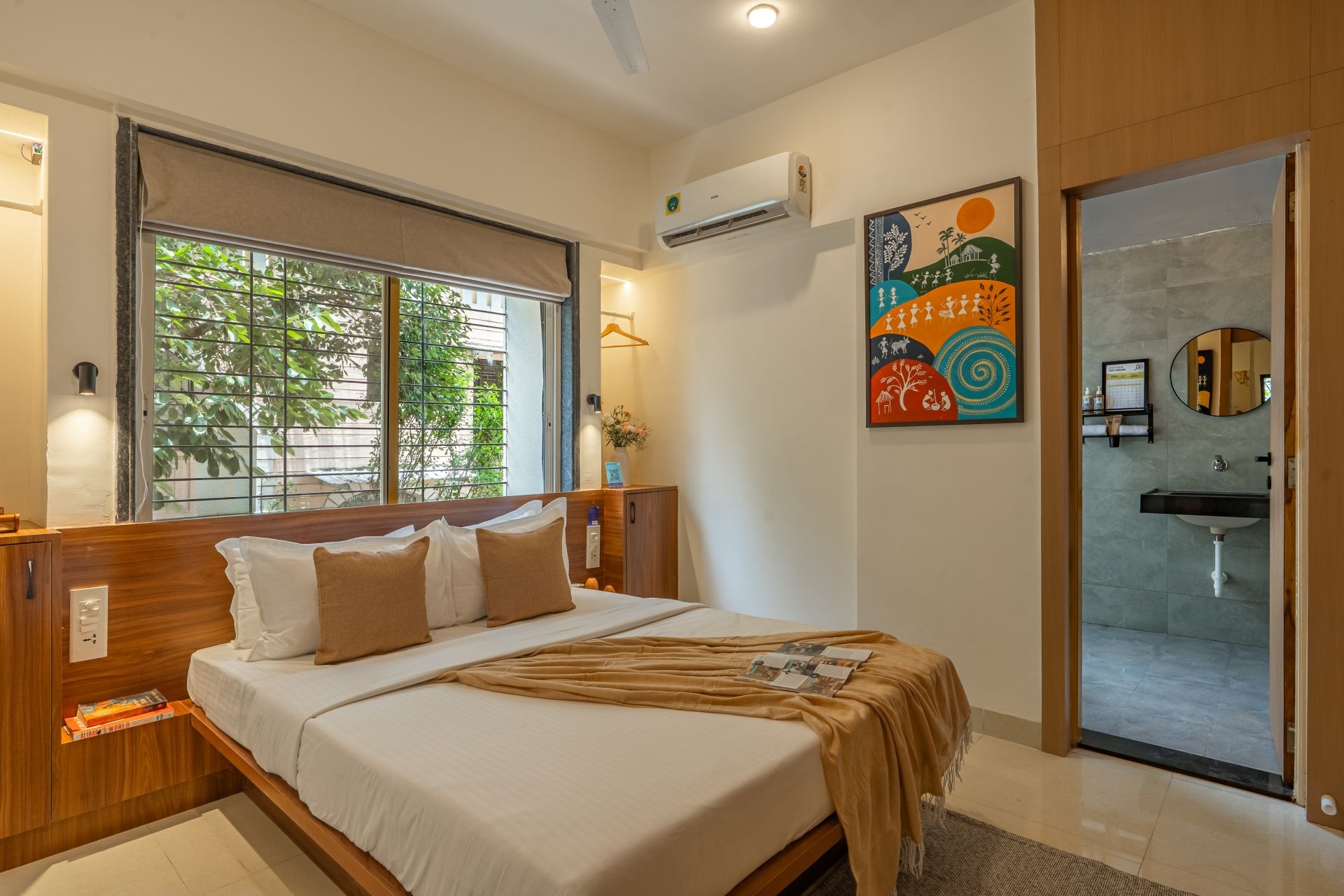 Habitación de hotel con cama, ventana con vistas a zonas verdes, unidad de aire acondicionado, una obra de arte colorida, espejo y baño con lavamanos.