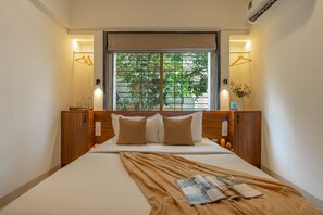 A hotel room with a bed, a window with a view of greenery, and a bedside table with a vase of flowers. - LiveAway Pune (Viman Nagar) by PACK (Pune)