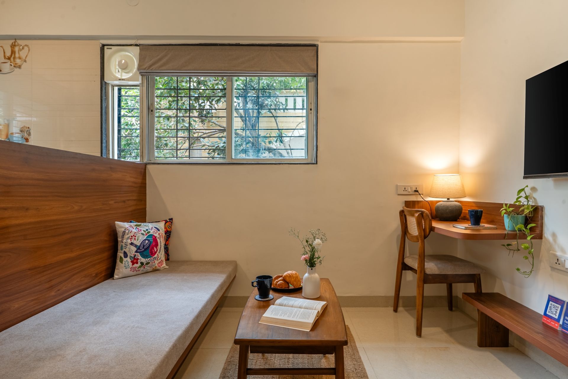 A cozy room with a wooden sofa, a small table with a book and a cup, a chair, a TV, and a window with a view of greenery.
