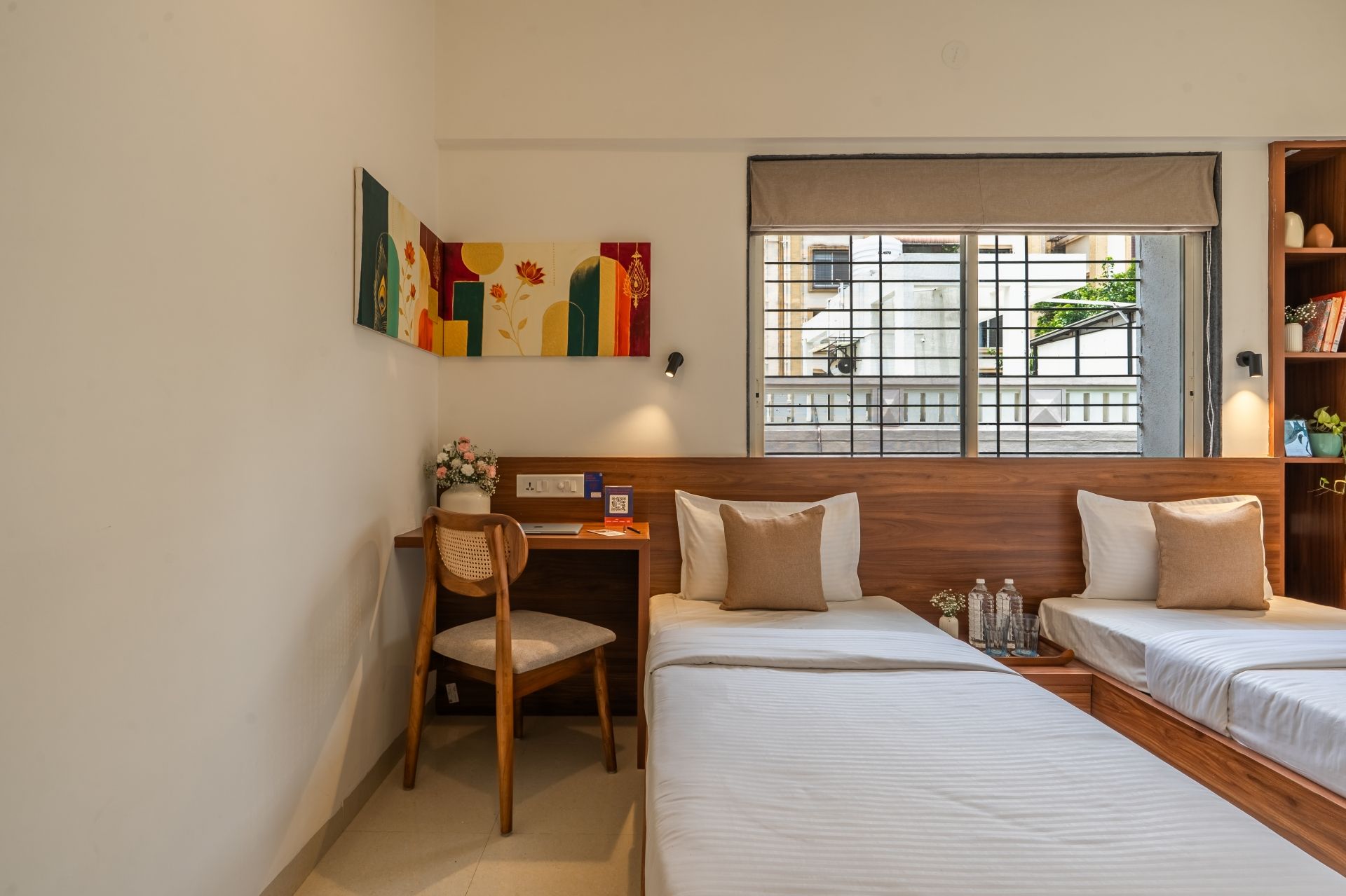 A hotel room with a desk, two beds, a chair, a window with a view of buildings, and a wall with artwork.