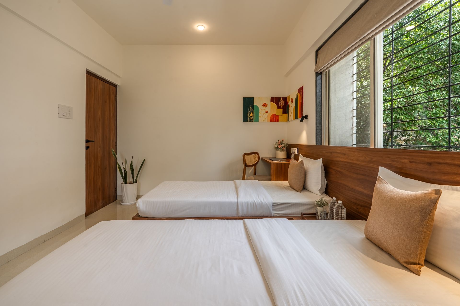 A bedroom with a large bed, a bedside table, a chair, a window with a view of greenery, and a door.