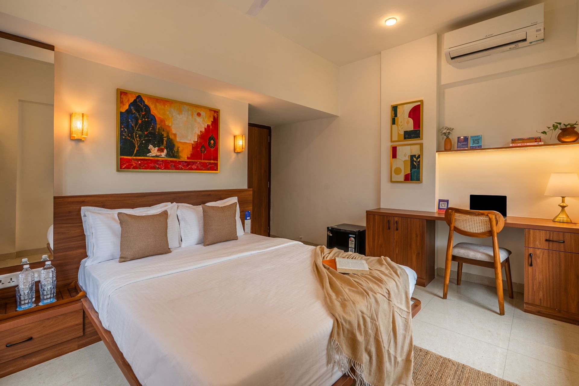 A hotel room with a bed, bedside table, desk, chair, and artwork on the walls.