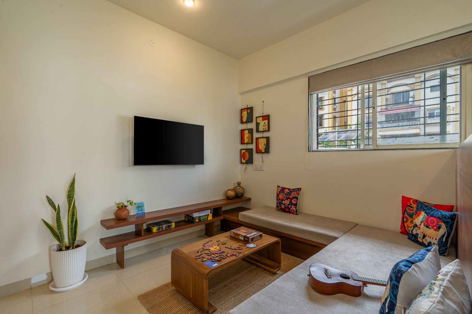 A living room with a flat-screen TV, a sofa, a coffee table with a puzzle, a guitar, and a window with a view of buildings.