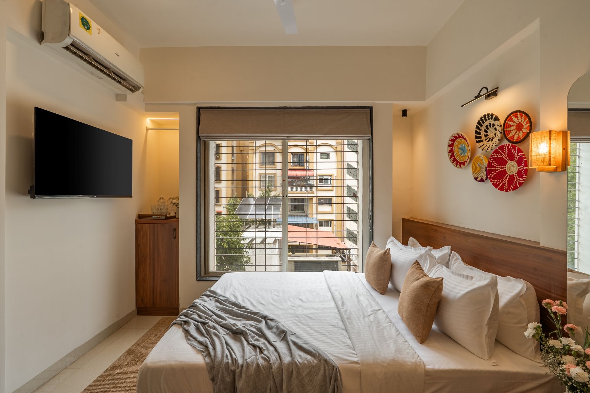 A bedroom with a large bed, a flat-screen TV mounted on the wall, a window with a view of buildings, and decorative wall art.