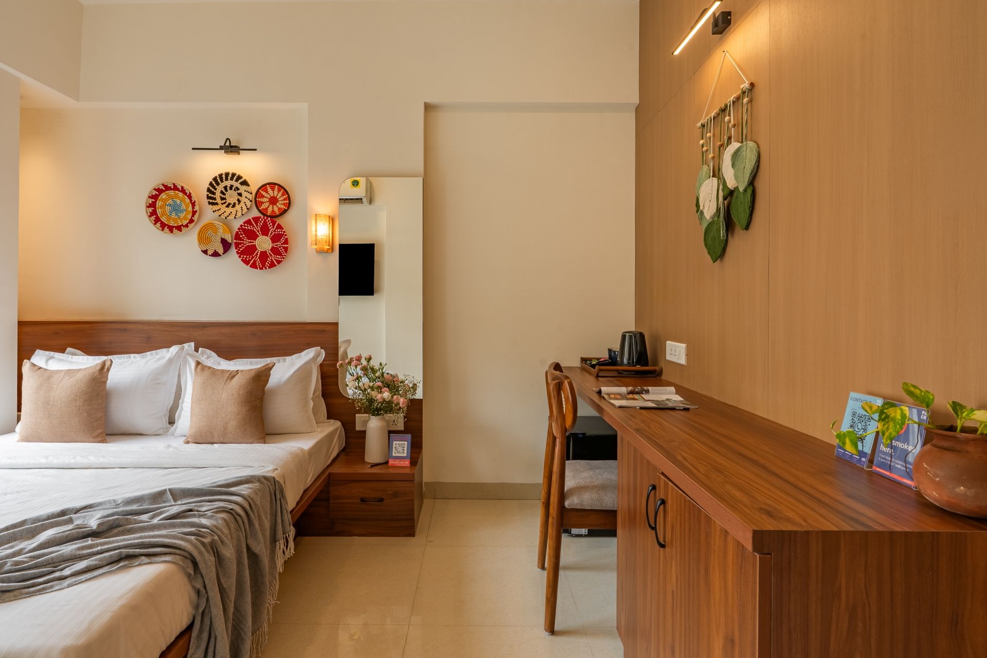 A hotel room with a bed, bedside table, and a desk with a chair. There are decorative wall hangings and a potted plant on the desk.