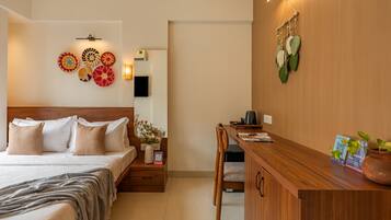 A hotel room with a bed, bedside table, and a desk with a chair. There are decorative wall hangings and a potted plant on the desk.