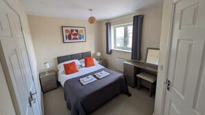 A bedroom with a bed, bedside table, lamp, desk, and a window with curtains. - Thistle Cottage (Bury St Edmunds)