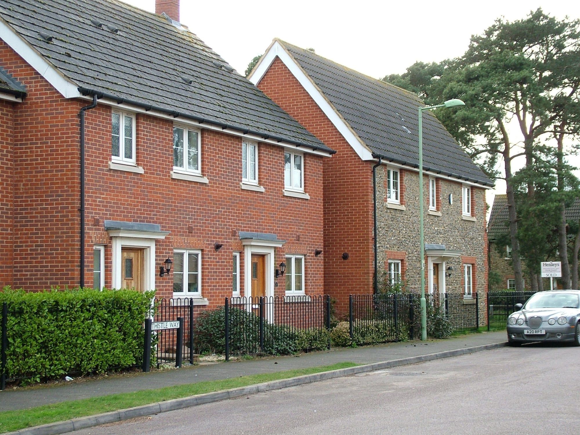 A row of two-story brick houses with pitched roofs, a metal fence, and a street sign indicating 'Thistle Way'.