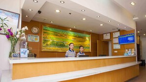 Un comptoir d’accueil avec deux employés, une horloge, un grand tableau représentant un paysage et divers affiches d’information.