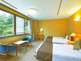A hotel room with a bed, a desk, a chair, a mirror, and a window with a view of greenery.
