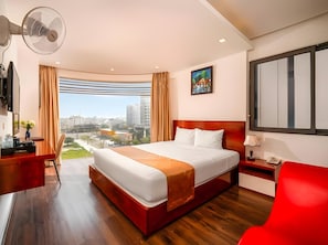 A hotel room with a large bed, a desk, a chair, a TV, and a view of the city. - Grand Maliya Hotel (Da Nang Municipality)