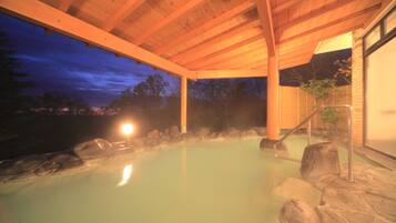 A hot spring area with a wooden roof structure, stone seating, and a view of the surrounding landscape.