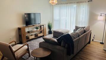 Ett vardagsrum med en soffa, en fåtölj, ett litet runt bord, en tv på ett träställ, en takfläkt och en golvlampa.