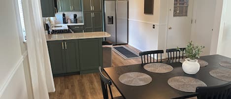 A modern kitchen with dark green cabinets, a stainless steel refrigerator, and a dining area with a dark wooden table and black chairs.