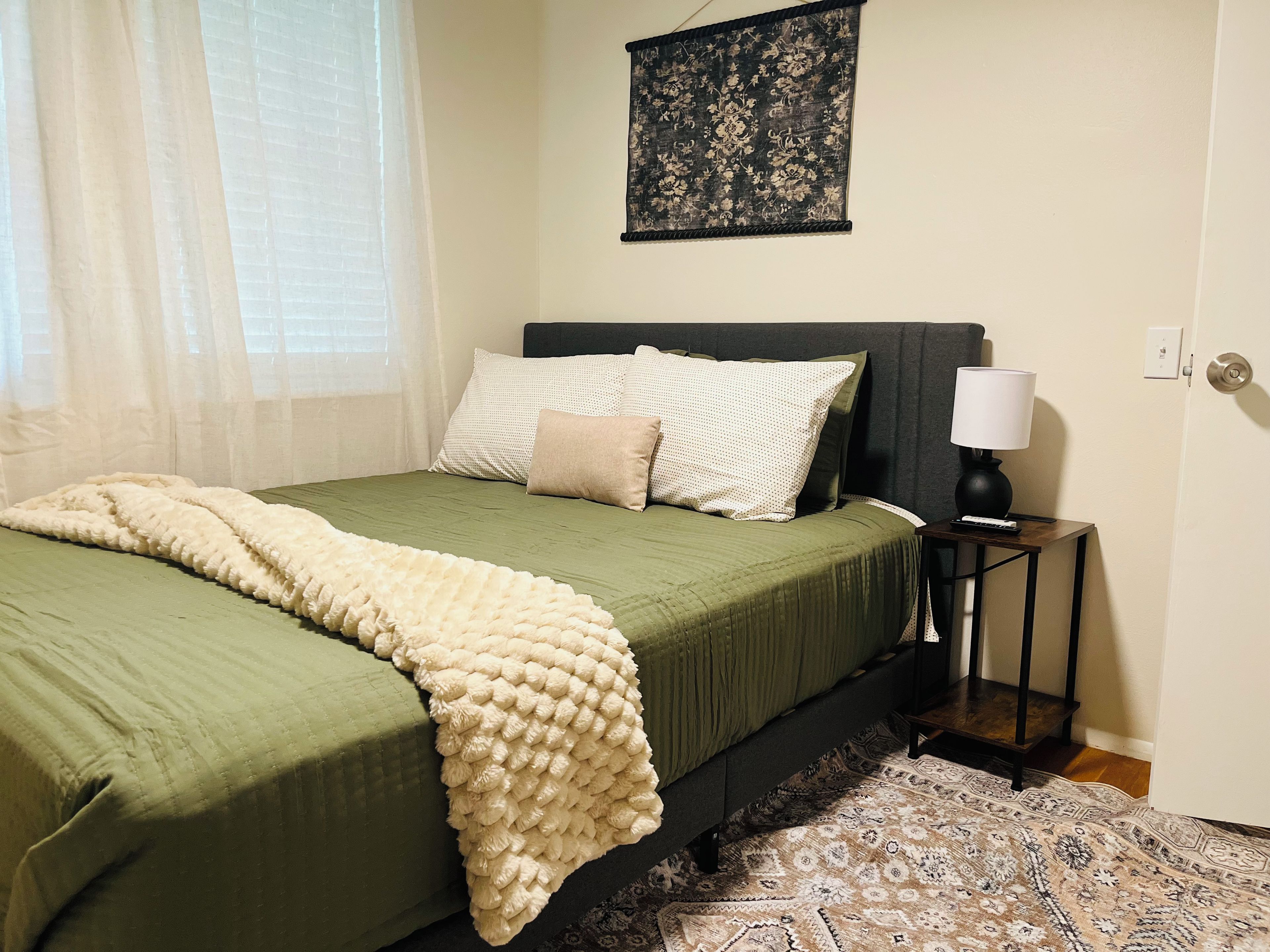 A neatly made bed with a green bedspread and a white throw blanket, a bedside table with a lamp, and a patterned rug on the floor.