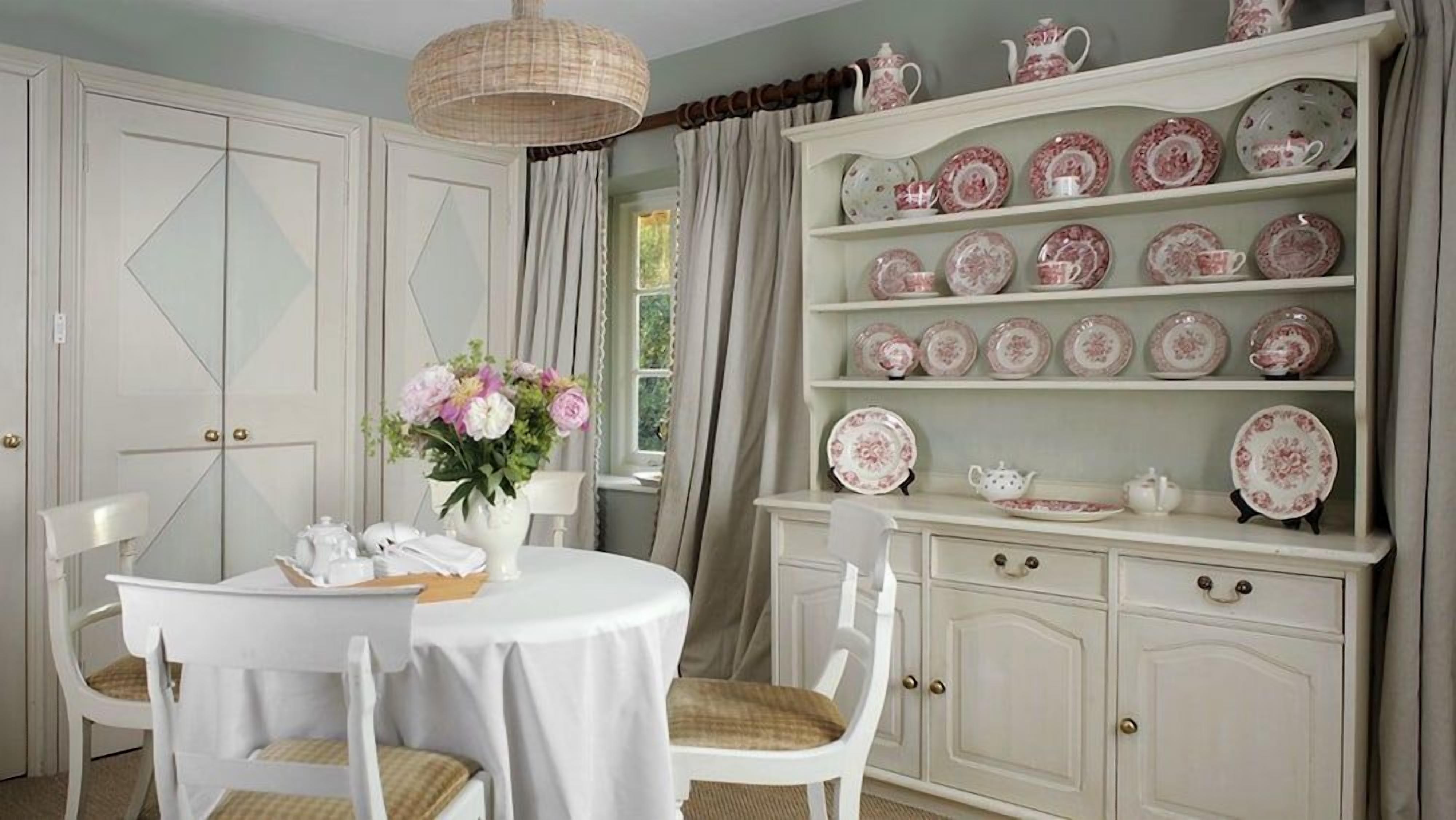 A dining area with a round table, white chairs, a vase of flowers, and a china cabinet with plates and cups.
