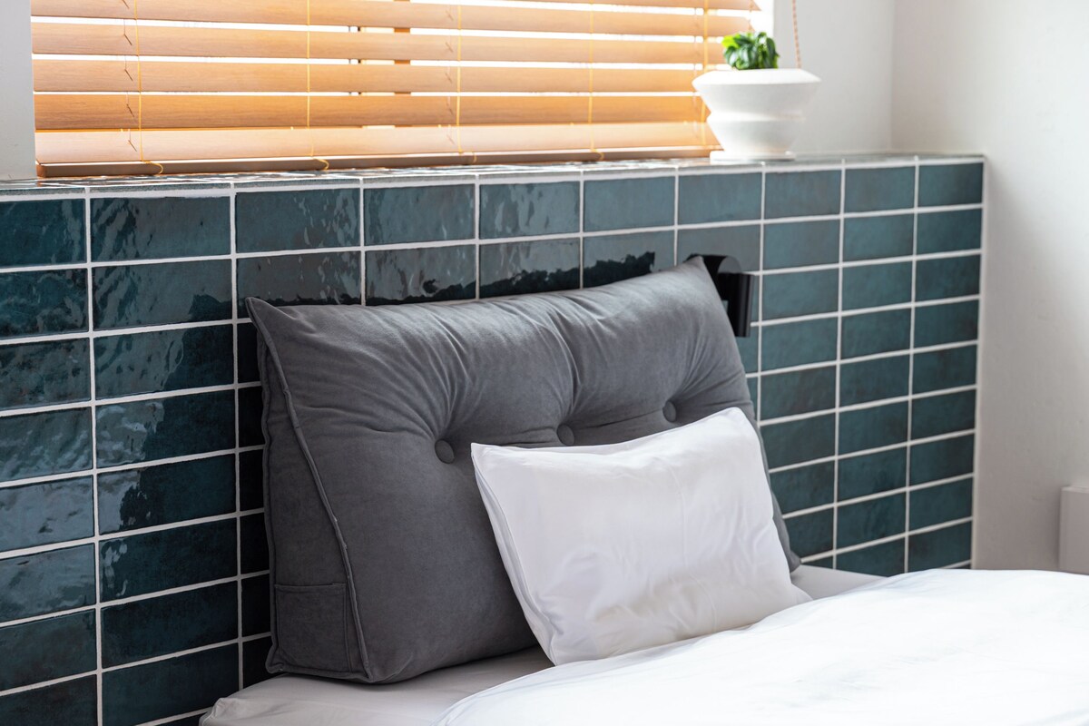 Ein Bett mit Kopfteil an einer gefliesten Wand, ein Fenster mit Jalousien und eine kleine Pflanze auf dem Fensterbrett.