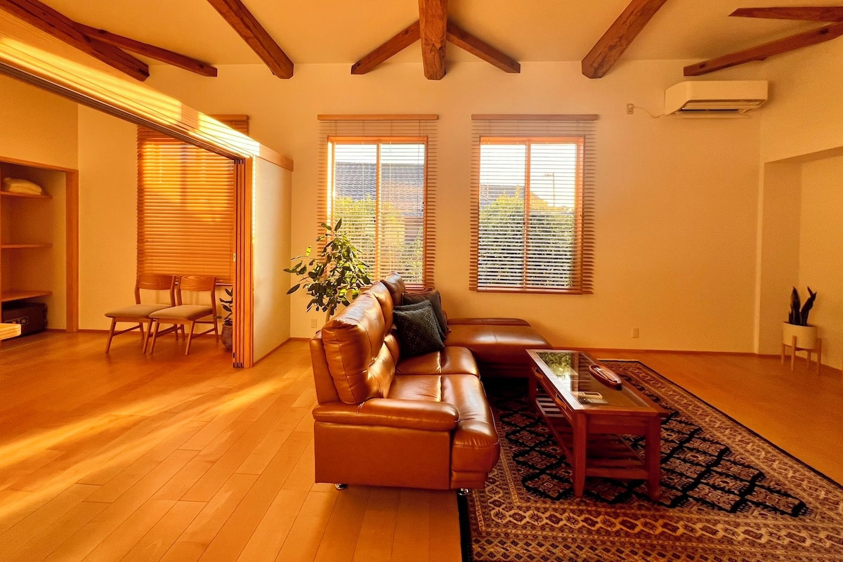 A spacious living room with a leather sofa, a coffee table, and a patterned rug. There are two windows with blinds, a wooden ceiling, and a built-in bookshelf.