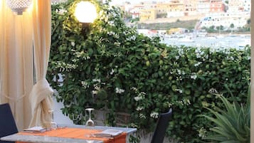 Ein Essbereich im Freien mit Tisch und Stühlen, einem Blumentopf und Blick auf eine Stadtkulisse durch eine Glastür.